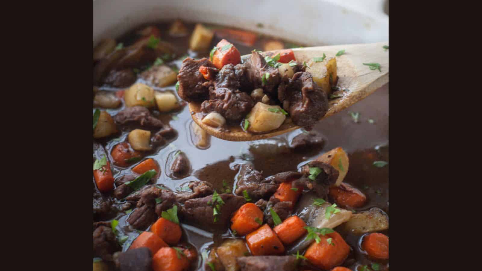 A wooden spoon holding beef stew with chunks of meat, carrots, and potatoes in a broth, garnished with herbs.