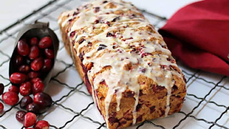 Loaf of cranberry bread with icing on a cooling rack, next to a scoop of fresh cranberries and a red napkin.