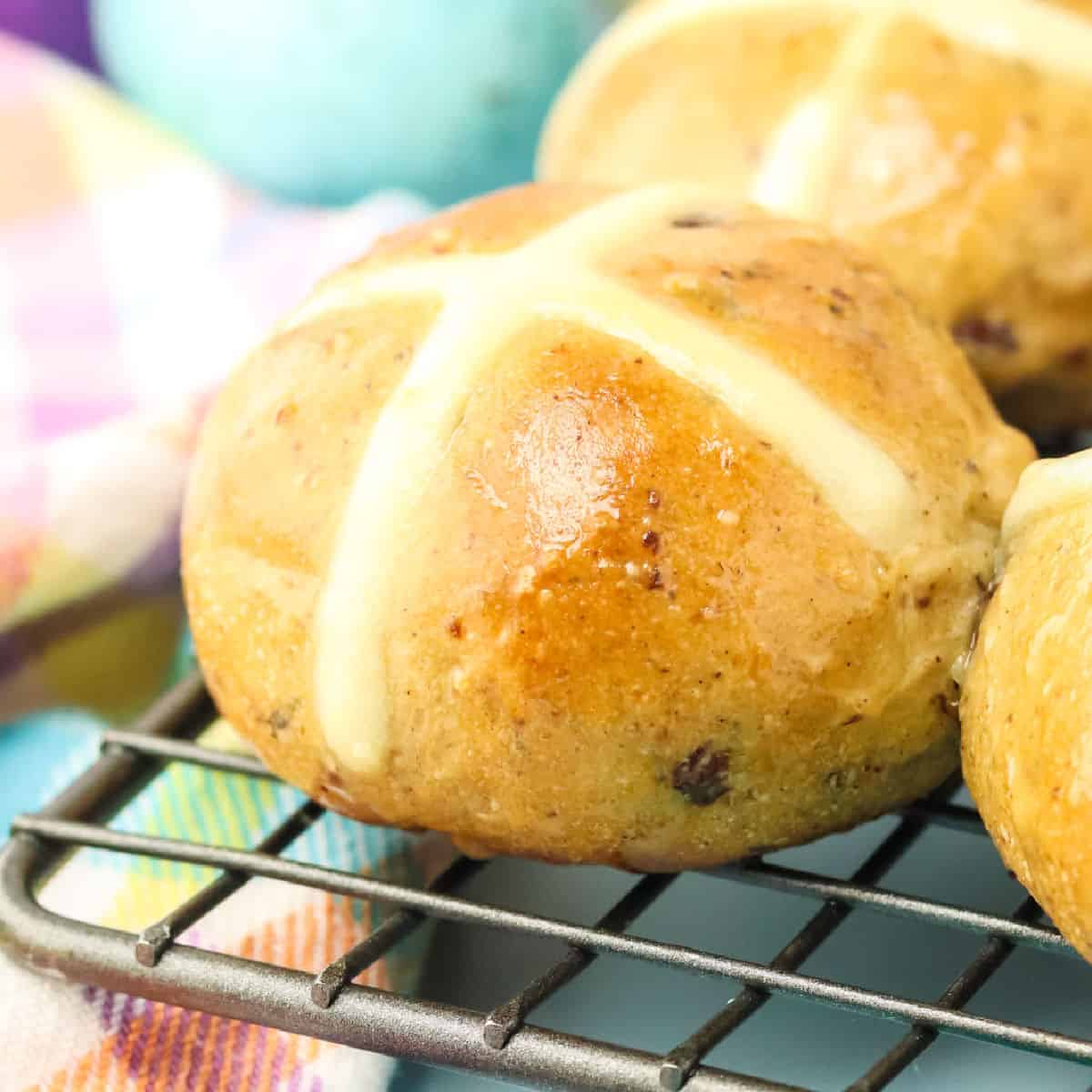 Close-up of baked hot cross buns with white icing crosses, resting on a wire rack. The background features colorful fabric and a blue egg.