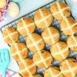 Hot cross buns cooling on a wire rack with Easter eggs and a spatula nearby on a light blue surface.