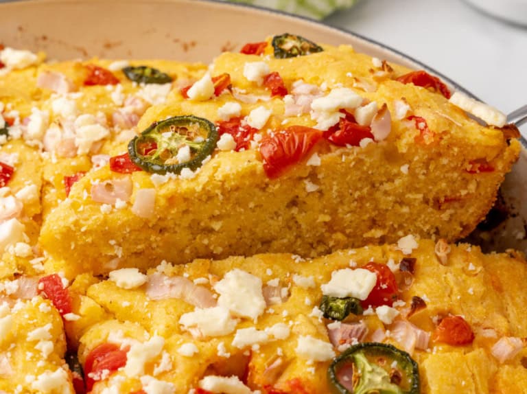 Close-up of cornbread topped with jalapeños, tomatoes, onions, and cheese, with a slice being lifted from the pan.