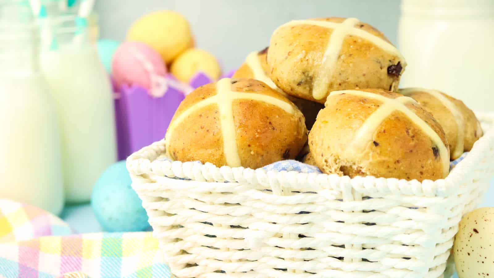 Basket of hot cross buns with a white cross on top, surrounded by pastel-colored eggs and bottles of milk.