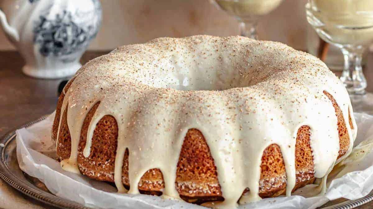 Bundt cake with white glaze drizzled over the top, placed on a tray. Background includes a teapot and two glass dishes.