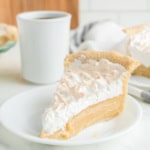 A slice of cream pie with a golden crust and whipped topping on a white plate, set next to a cup of black coffee.