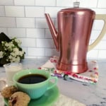 Copper coffee pot on a floral cloth, a green cup of coffee with cookies, glass of milk, and a small bouquet of white flowers on a tiled background.