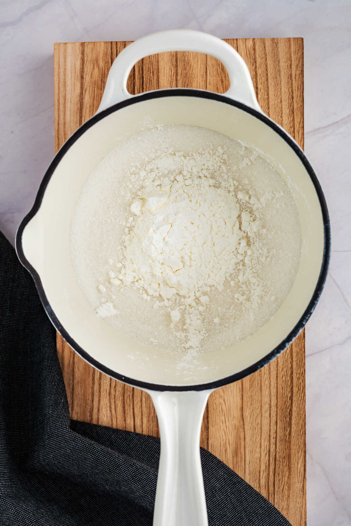 A white saucepan containing a mixture of flour and sugar sits on a wooden board. A dark cloth is partially visible beside it.