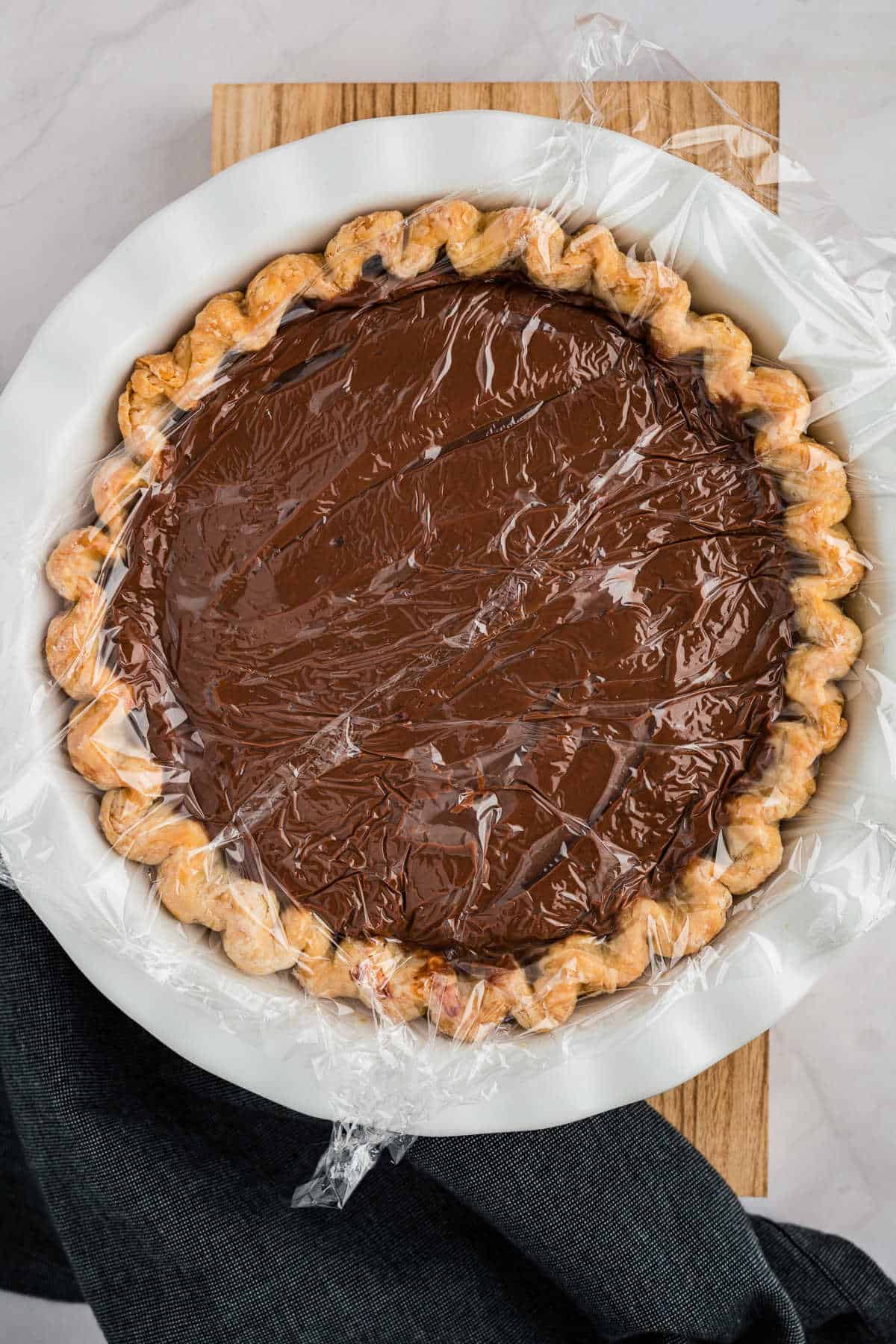 A chocolate cream pie topped with plastic wrap in a white pie dish on a wooden board.