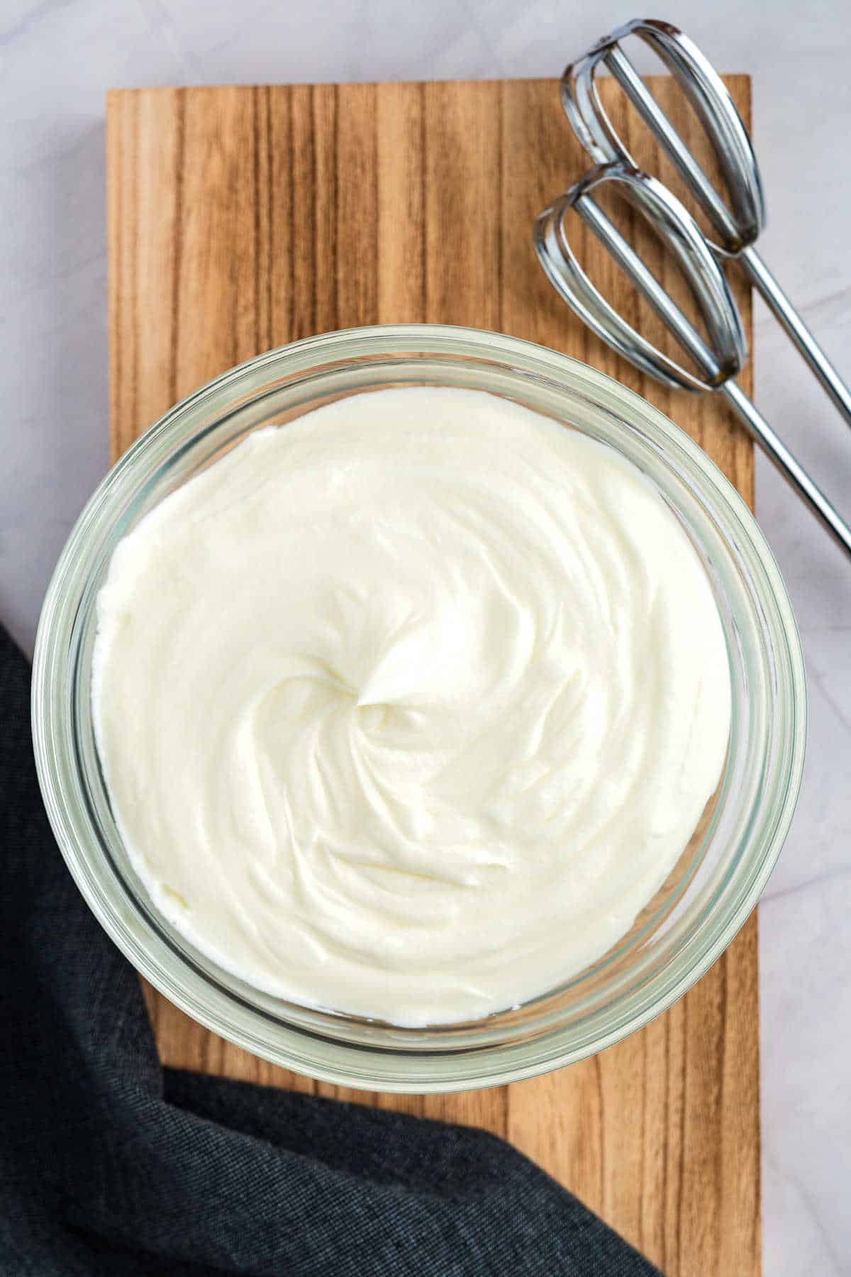 A bowl of smooth, whipped cream on a wooden board, with a pair of metal beaters and a dark cloth visible nearby.