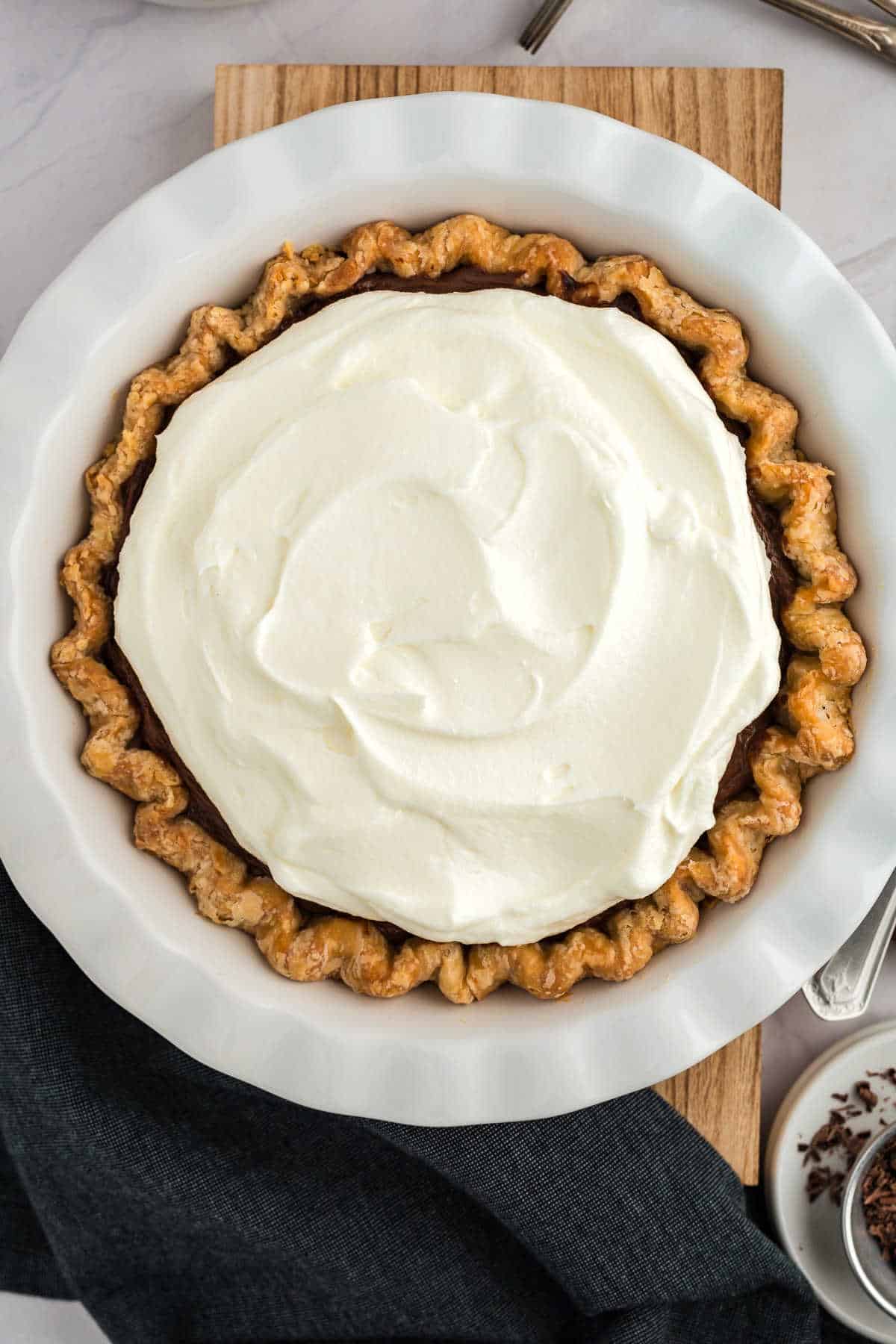 A pie with a golden crust, topped with a layer of smooth whipped cream, sits on a wooden board. A dark napkin and a small bowl with a spoon are nearby.