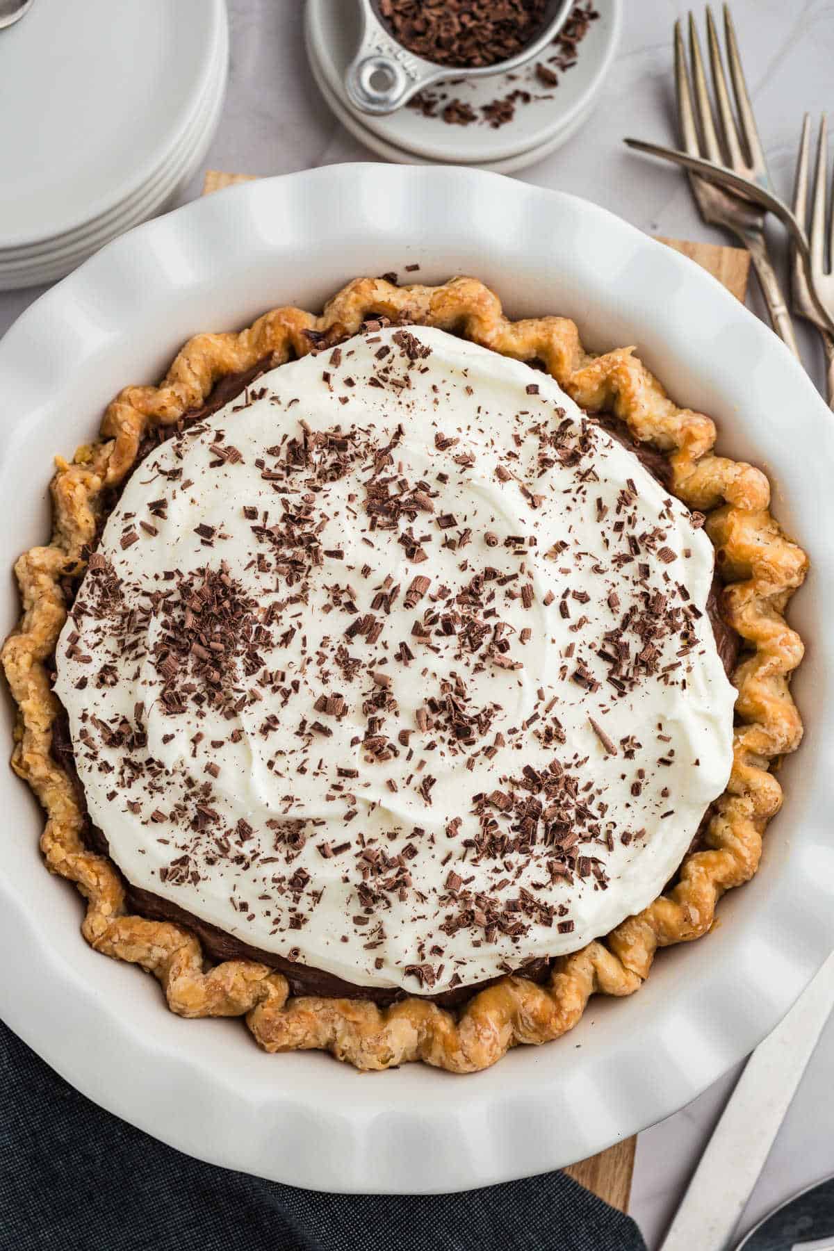 A pie with a crimped crust is topped with whipped cream and sprinkled with chocolate shavings, placed in a white dish. Plates and forks are nearby.