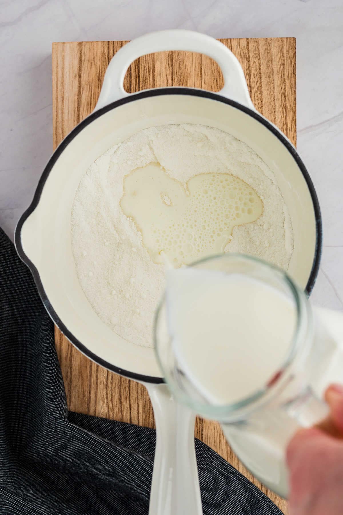 Milk is being poured from a jug into a white pot containing flour and oil, placed on a wooden board with a dark cloth beside it.