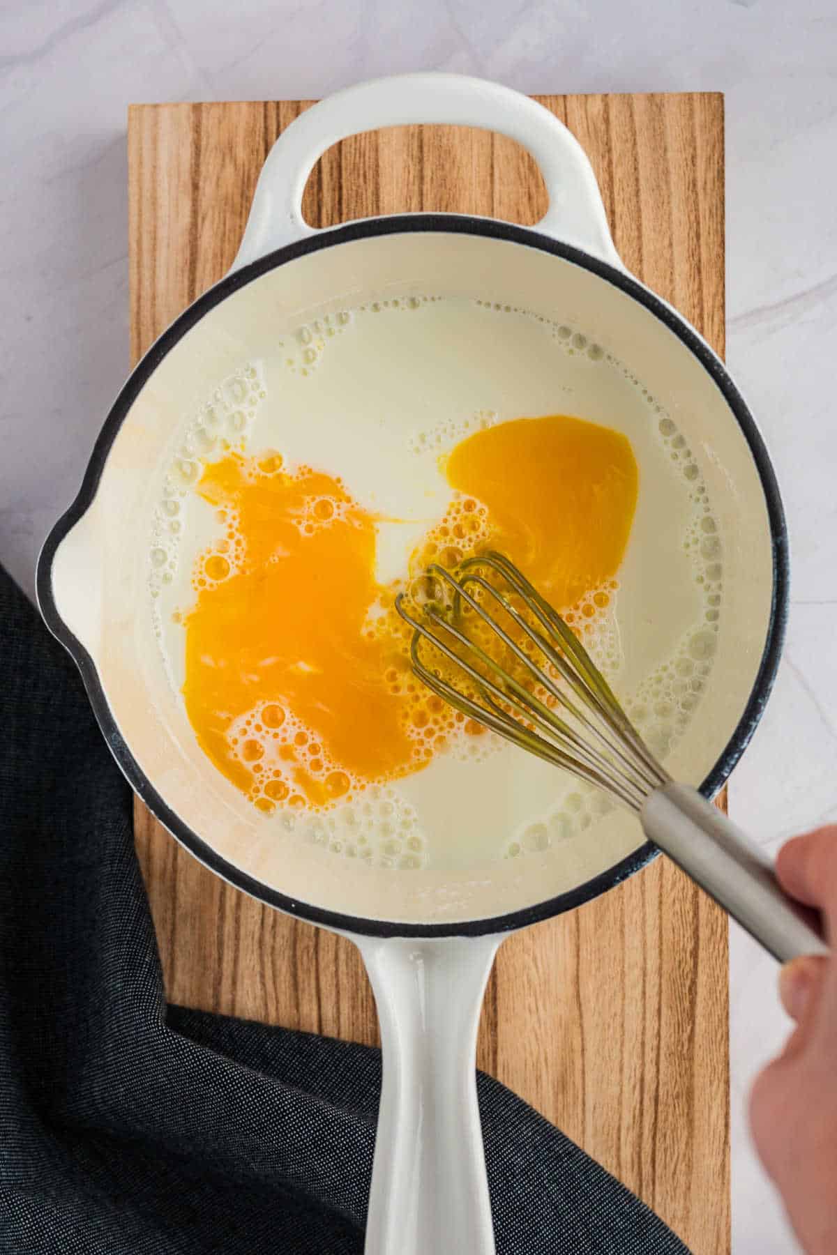 A hand whisking eggs in a white saucepan on a wooden cutting board.