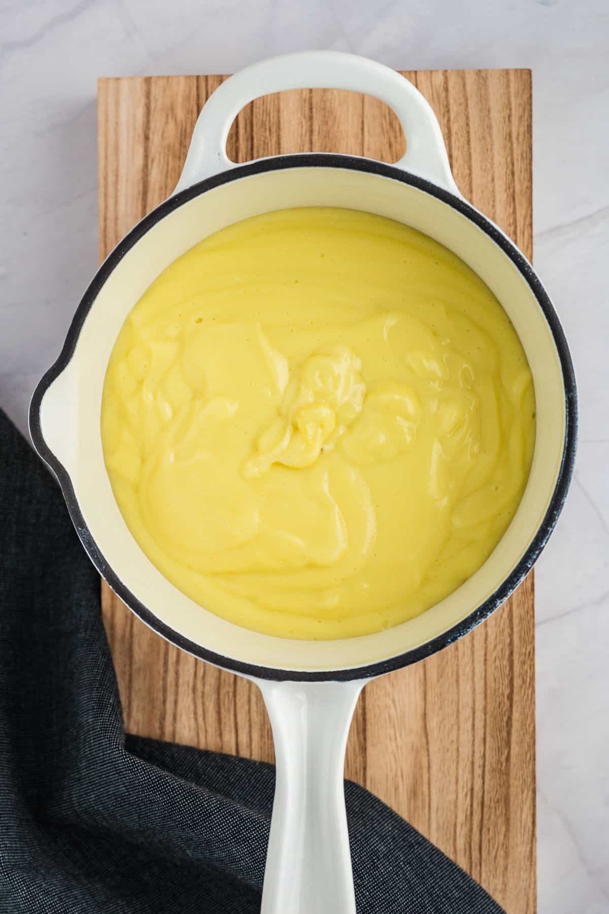 A white pot filled with thick, creamy yellow custard sits on a wooden board. A dark cloth is partially visible next to the pot.