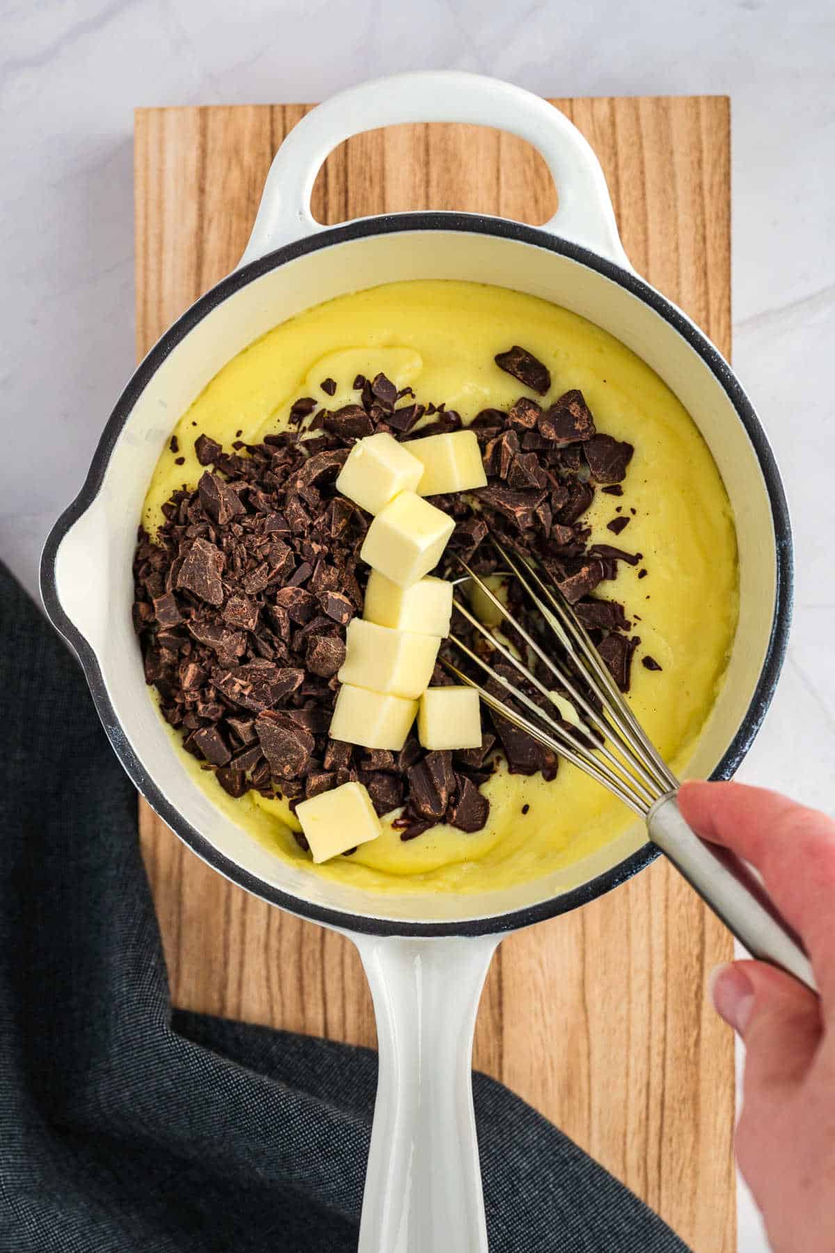 A person whisks cubed butter and chopped chocolate into a yellow mixture in a white saucepan on a wooden board.
