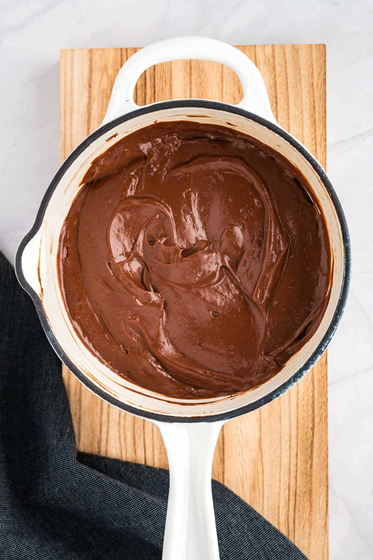 A white pot filled with smooth, creamy chocolate pudding resting on a wooden surface, with a dark cloth partially visible on the side.
