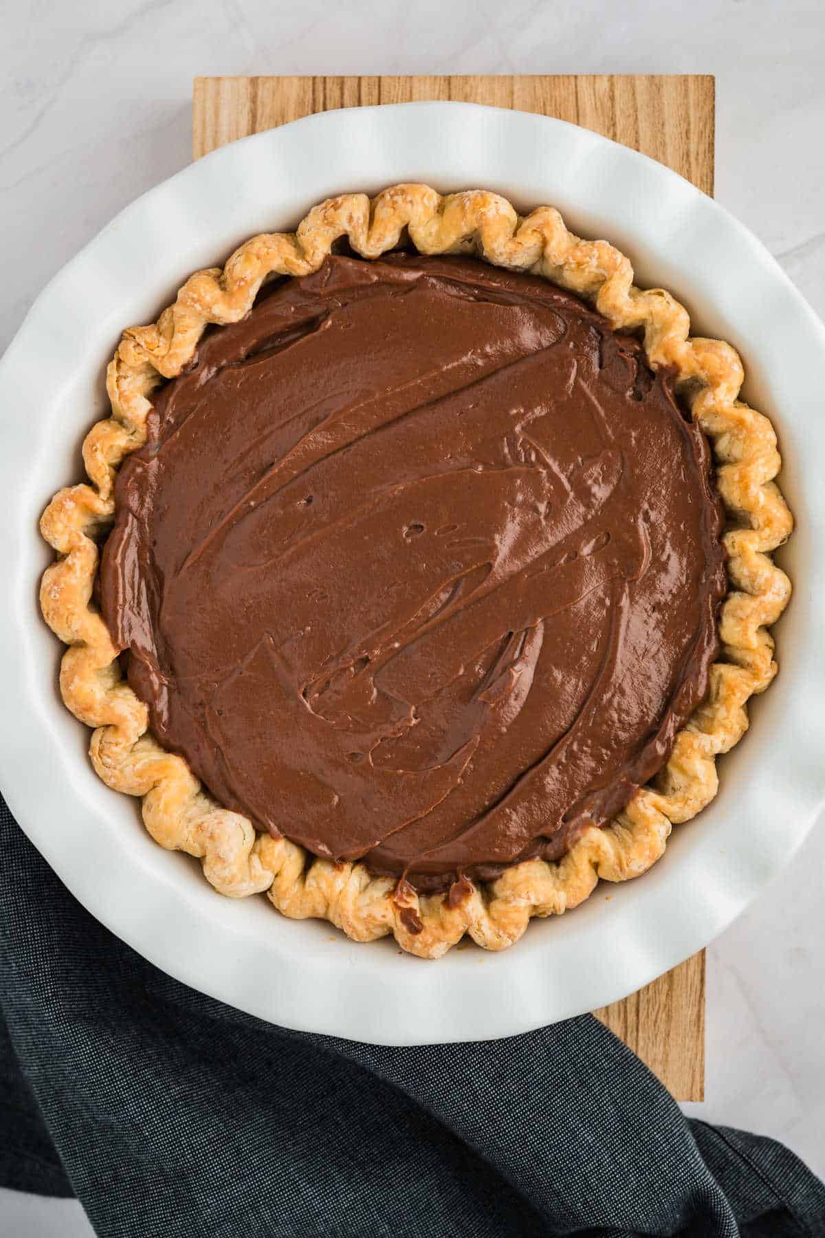 A chocolate pie with a wavy crust sits in a white dish on a wooden surface, partially covered by a dark cloth.