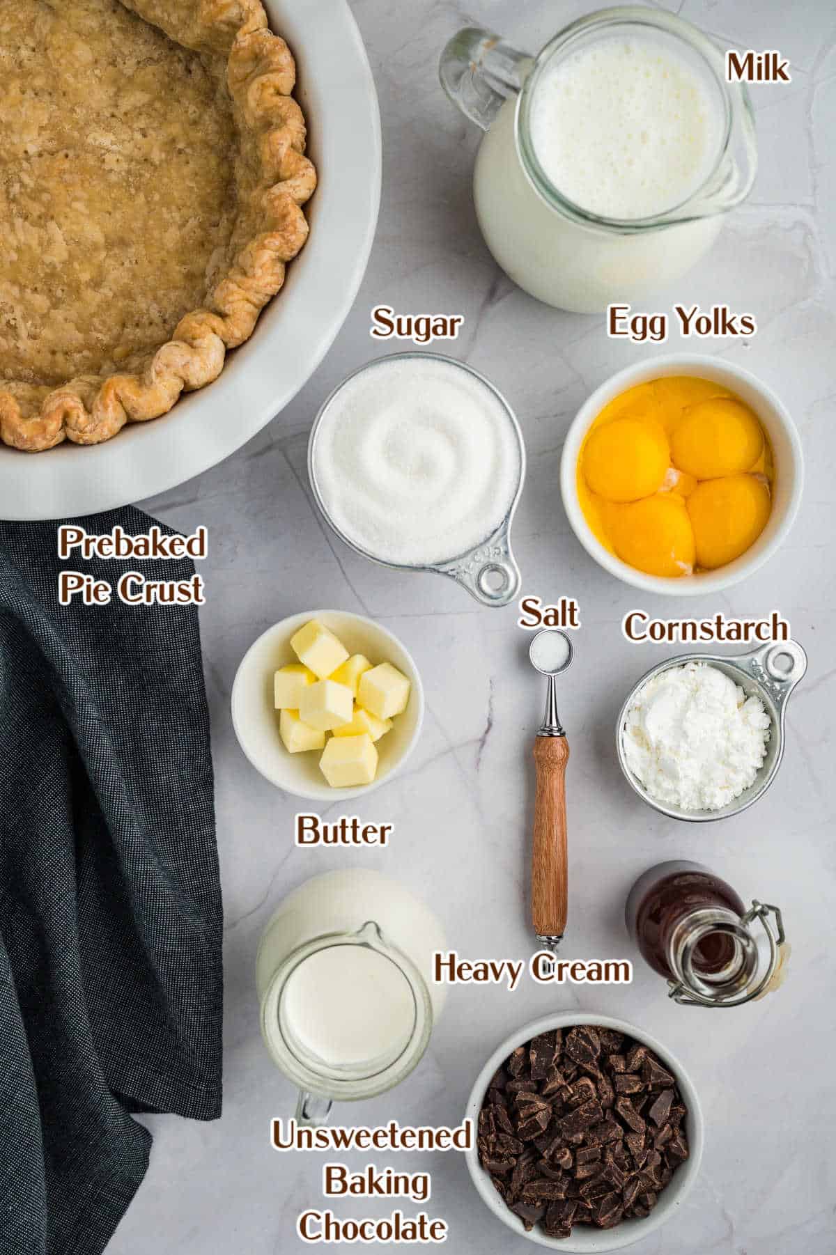Image of ingredients for a chocolate pie recipe laid out on a table. Includes a prebaked pie crust, milk, sugar, egg yolks, butter, salt, cornstarch, heavy cream, unsweetened baking chocolate.