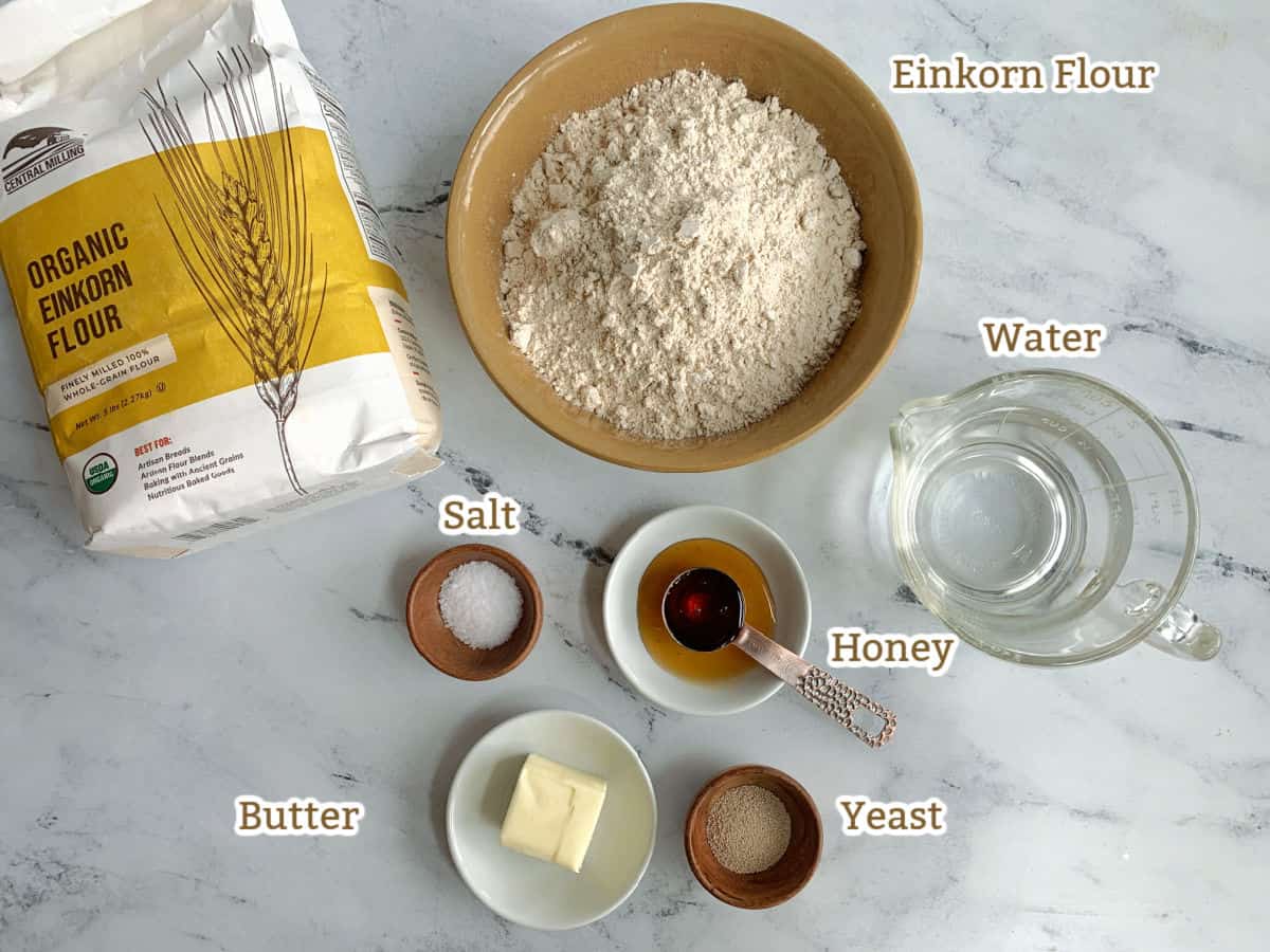 Einkorn bread ingredients on a marble surface: einkorn flour in a bowl and a bag, water, honey, yeast, salt, and butter.