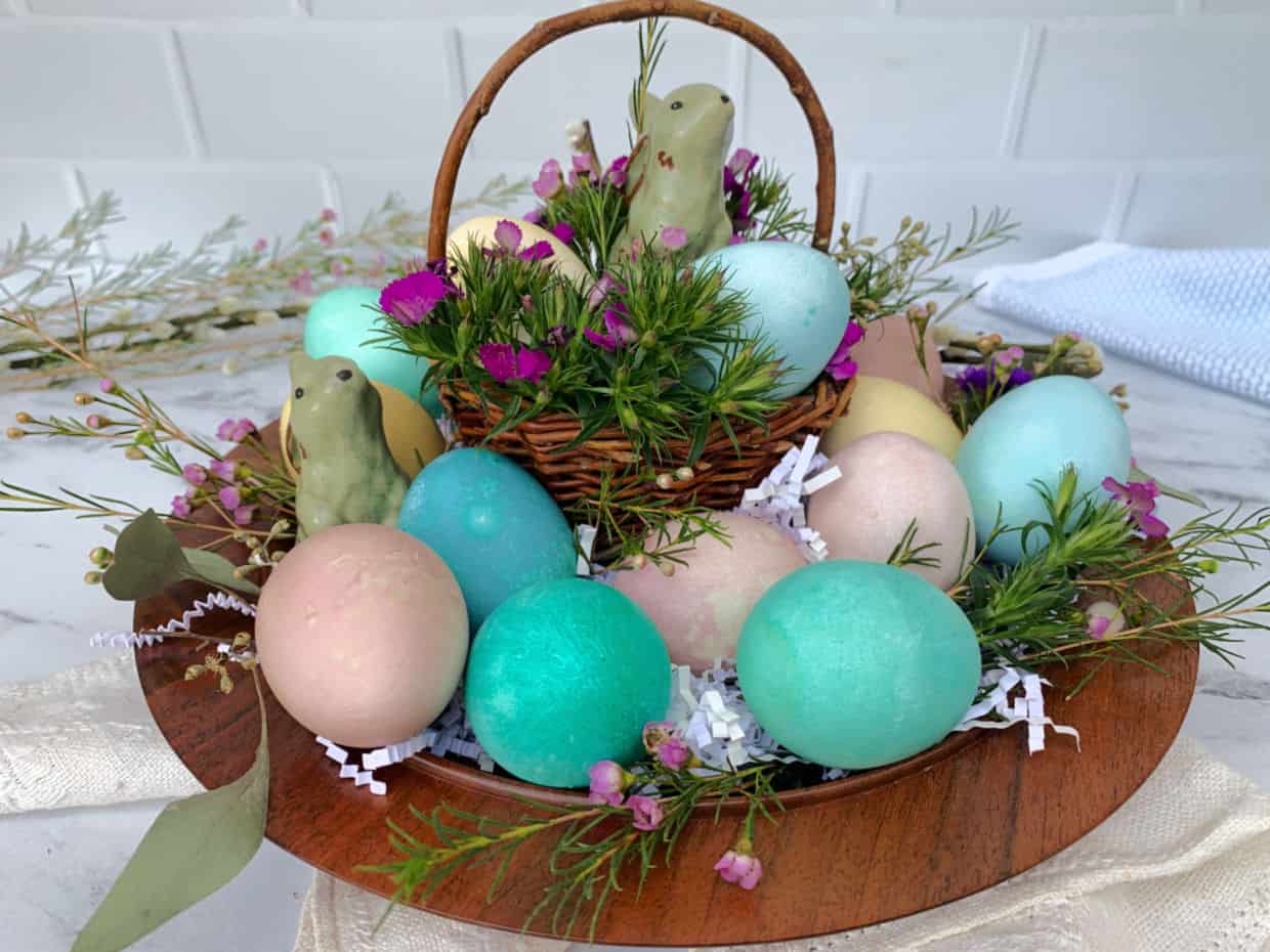 A wooden tray displays colorful pastel eggs, greenery, pink flowers, and bird figurines around a small woven basket on a marble surface.