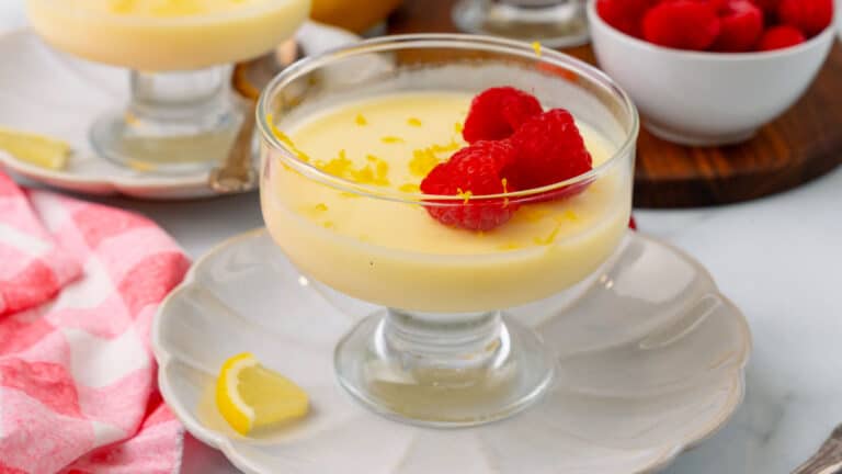 A glass dish of lemon pudding topped with fresh raspberries and lemon zest, placed on a white plate with a lemon wedge nearby. A bowl of raspberries is in the background.