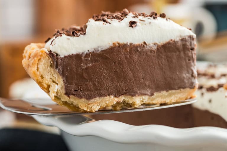 A slice of chocolate cream pie with whipped cream topping and chocolate shavings, served on a metal spatula over a white pie dish.