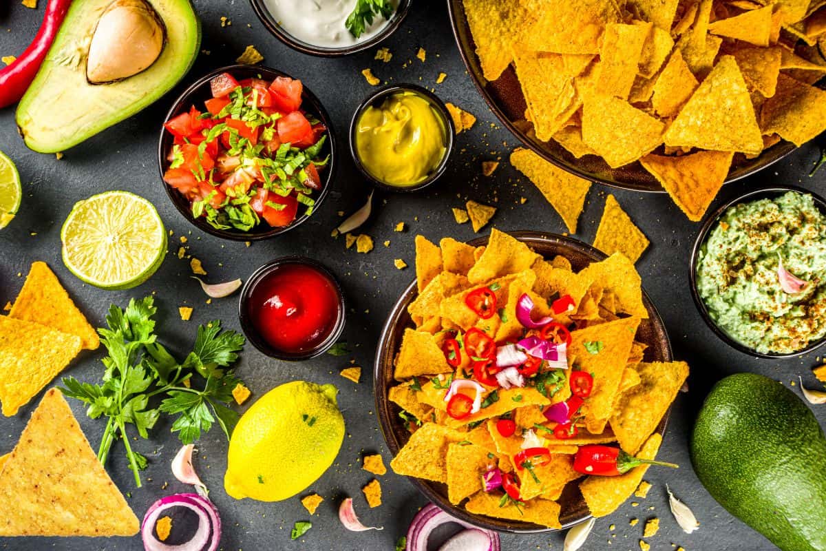 A creative spread of nachos featuring avocado, lime, salsa, onion, and parsley is laid out on a dark surface. This display offers delicious nacho bar ideas with an array of sauces and crispy tortilla chips.