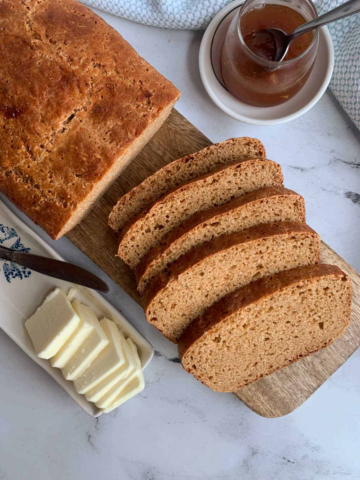 A loaf of Einkorn bread partially sliced on a wooden board, with a dish of butter slices and a jar of jam with a spoon beside it on a marble surface.