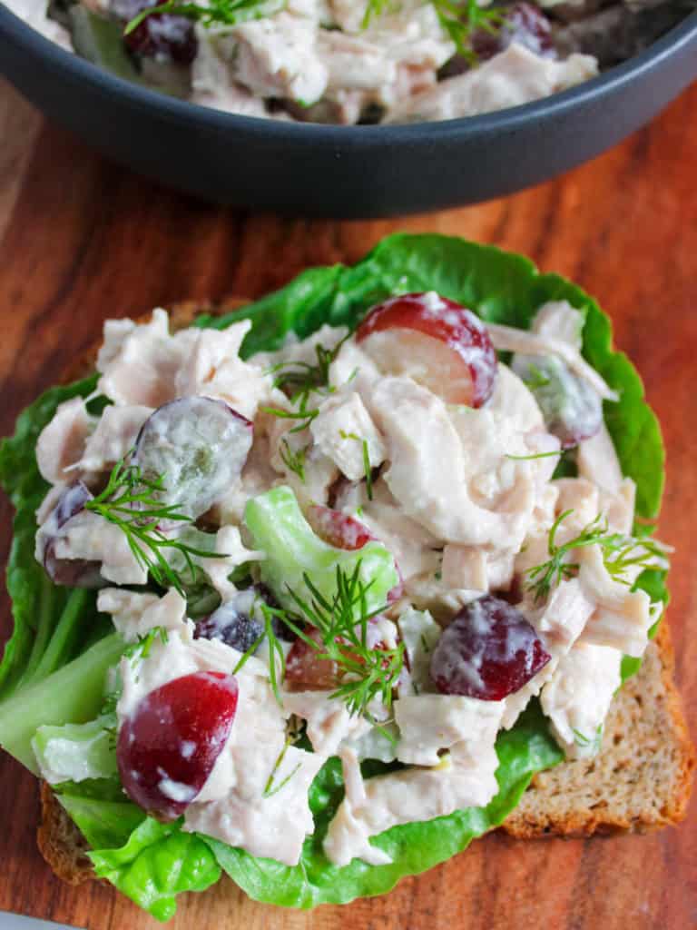 Open-faced chicken salad sandwich with grapes and fresh dill on lettuce, served on whole-grain bread, placed on a wooden surface.