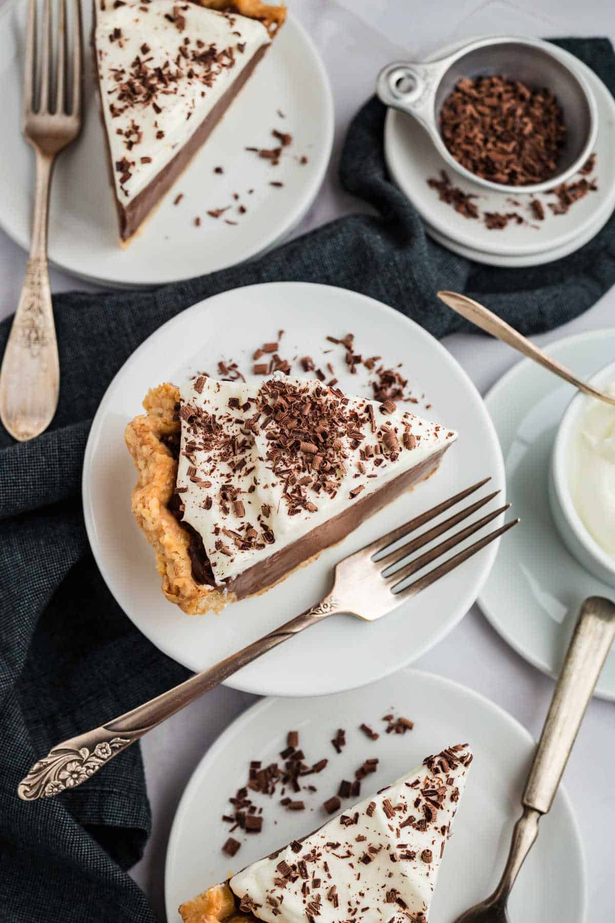 Slices of chocolate cream pie with whipped topping and chocolate shavings on white plates, with forks on a dark cloth.