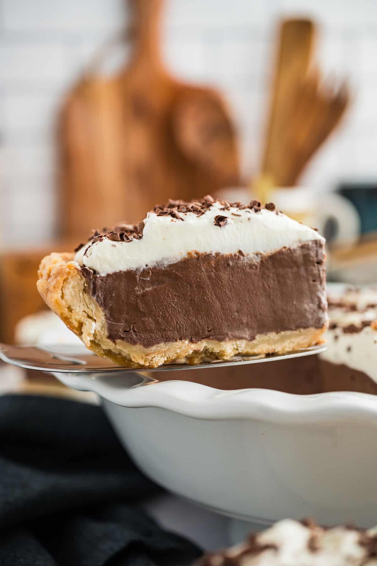 A slice of chocolate cream pie with whipped topping and chocolate shavings is being served from a white pie dish.