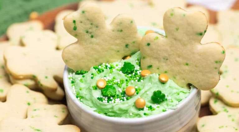 A bowl of green frosting topped with clover-shaped sprinkles, surrounded by shamrock-shaped cookies.