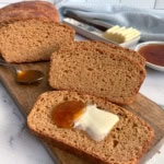 Sliced brown bread on a wooden board with one slice topped with jam and butter. A spoon with jam, butter slices, and a small dish of jam in the background.