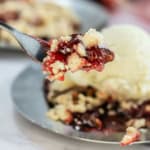 A fork holds a bite of berry crumble with red sauce, crumbs, and a scoop of vanilla ice cream on a metal plate.