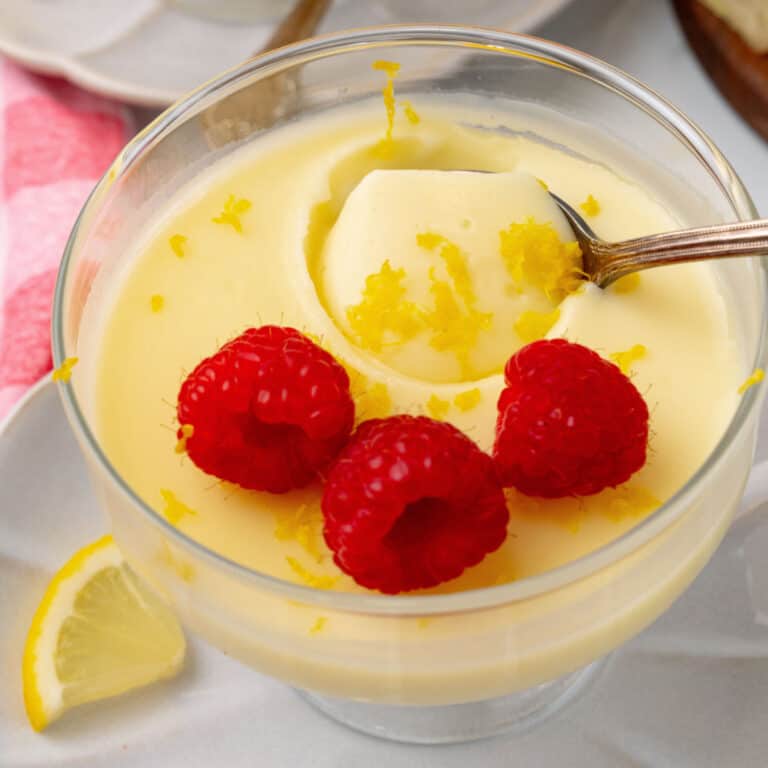 A glass bowl of lemon pudding topped with lemon zest and three raspberries, with a spoon in the pudding. A lemon wedge is on the side.