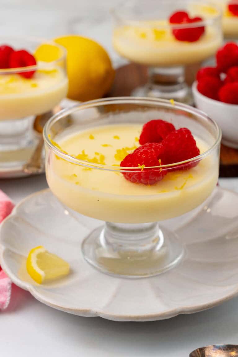Glass dessert dish with creamy lemon mousse, topped with raspberries and lemon zest. In the background, more dessert bowls, a lemon, and a bowl of raspberries are visible.