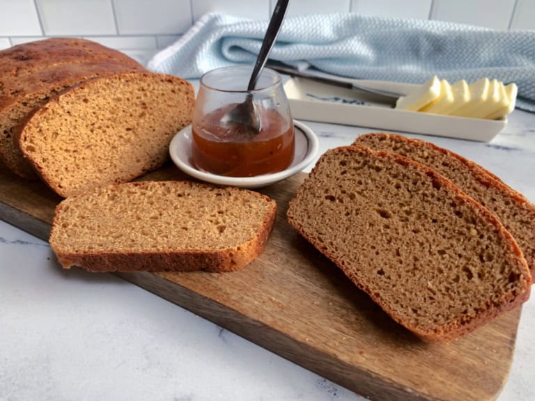 Sliced brown bread on a wooden board with a jar of jam and a butter dish in the background.