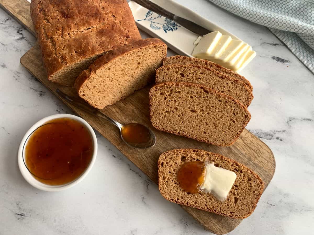 Sliced brown bread on a wooden board with butter and a small dish of marmalade, placed on a marble surface.