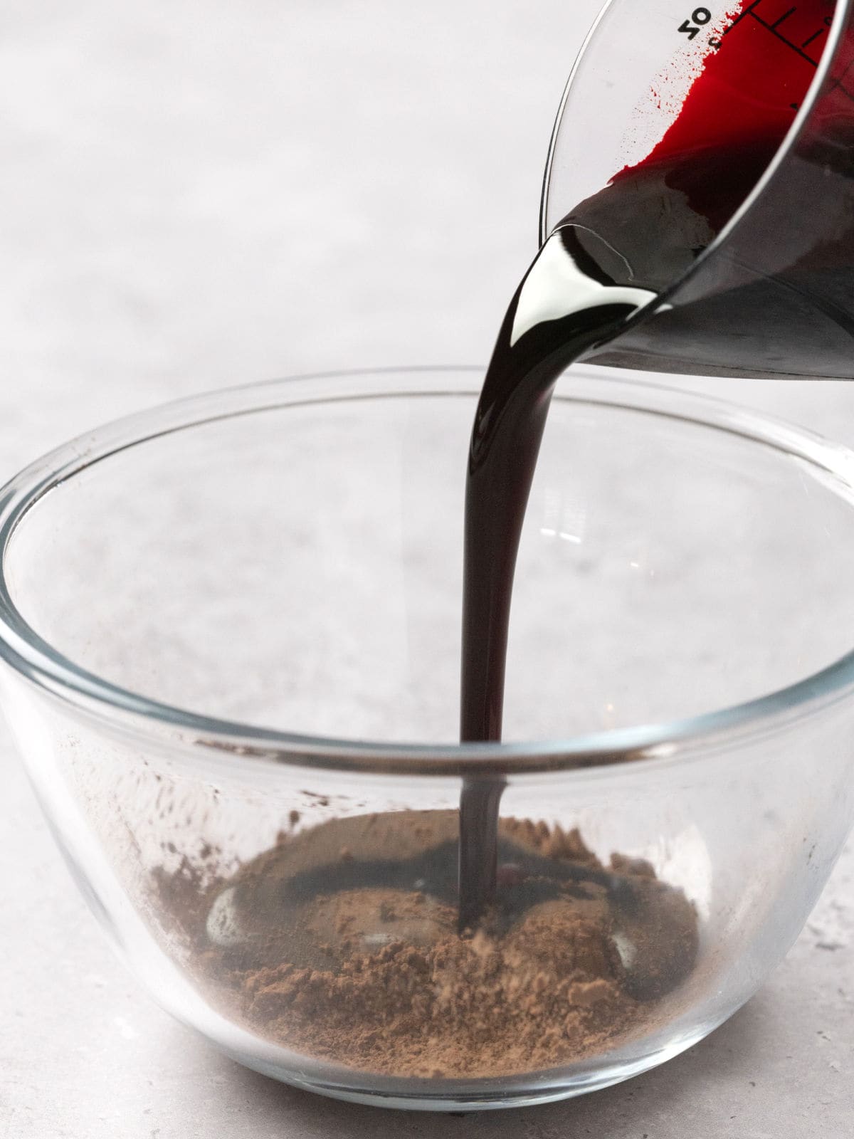 Pouring dark liquid into a clear glass bowl with powder, on a light gray surface.