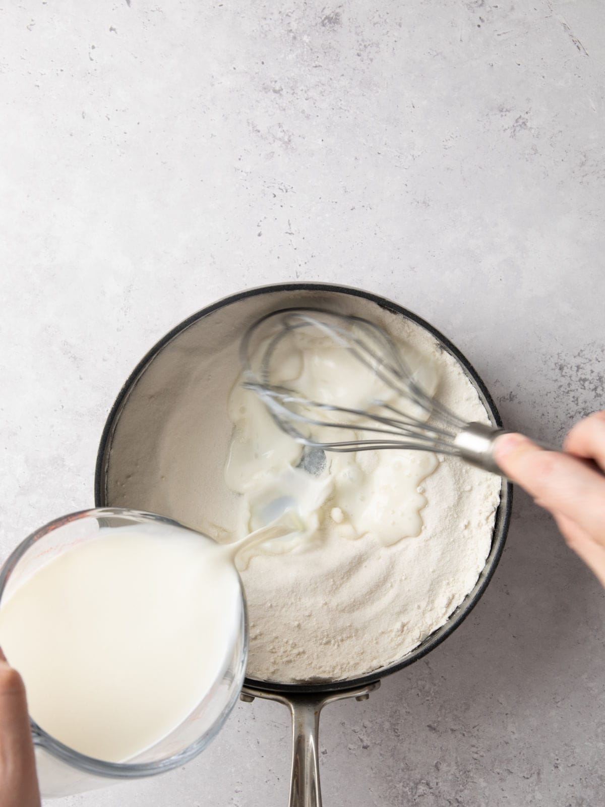 Close-up of a person whisking milk into flour in a saucepan on a gray surface.