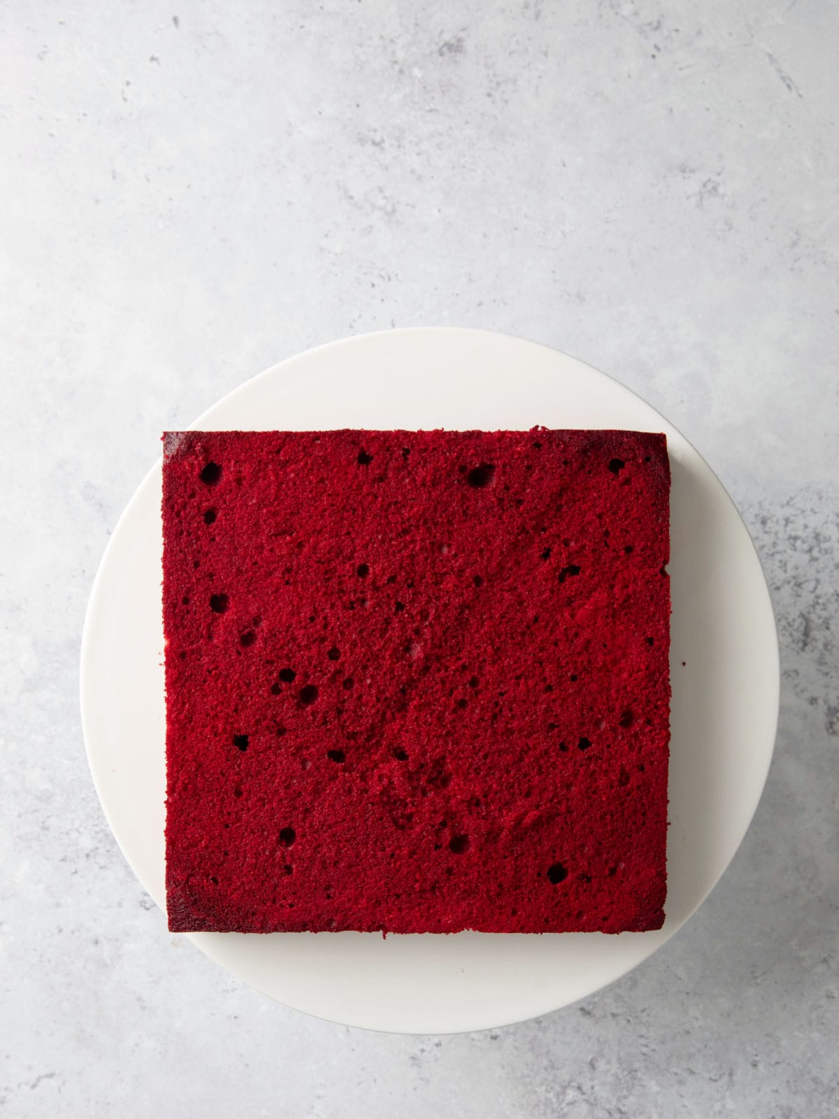 A square red velvet cake on a white plate, placed on a light marble surface.