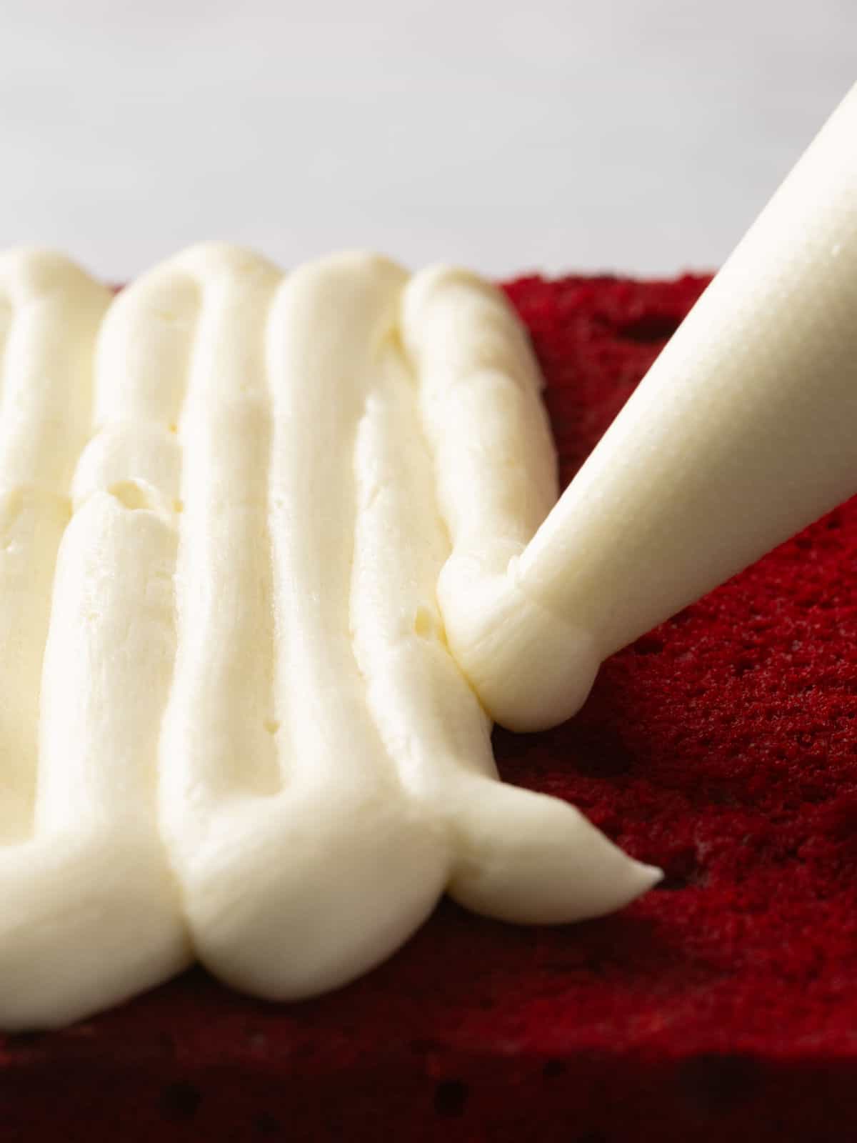 A piping bag applies creamy frosting in parallel lines onto a piece of red velvet cake.