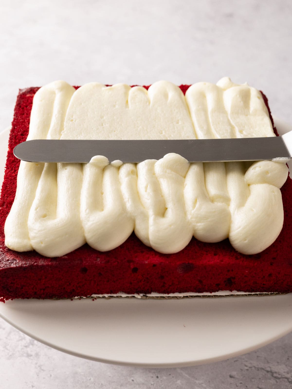 Frosting being evenly spread on a rectangular red velvet cake layer using an offset spatula on a white plate.