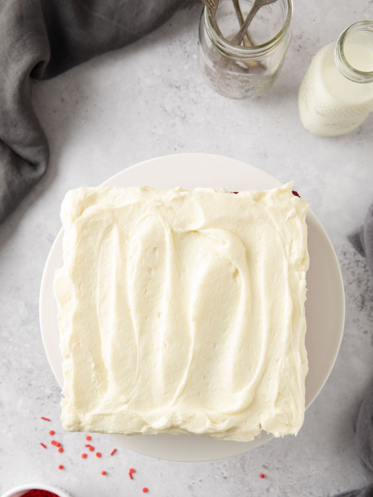 Square cake with smooth white frosting on a round white plate, surrounded by a glass bottle, a jar with straws, and a gray cloth on a light gray surface.