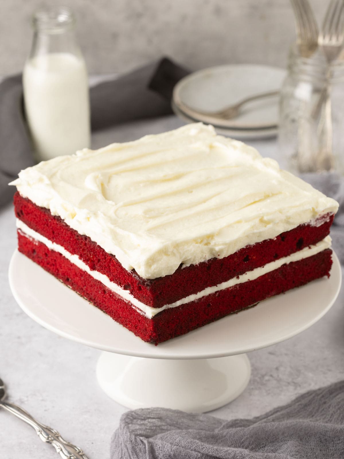 A rectangular red velvet cake with white frosting layers on a white cake stand. A bottle of milk and plates are in the background.