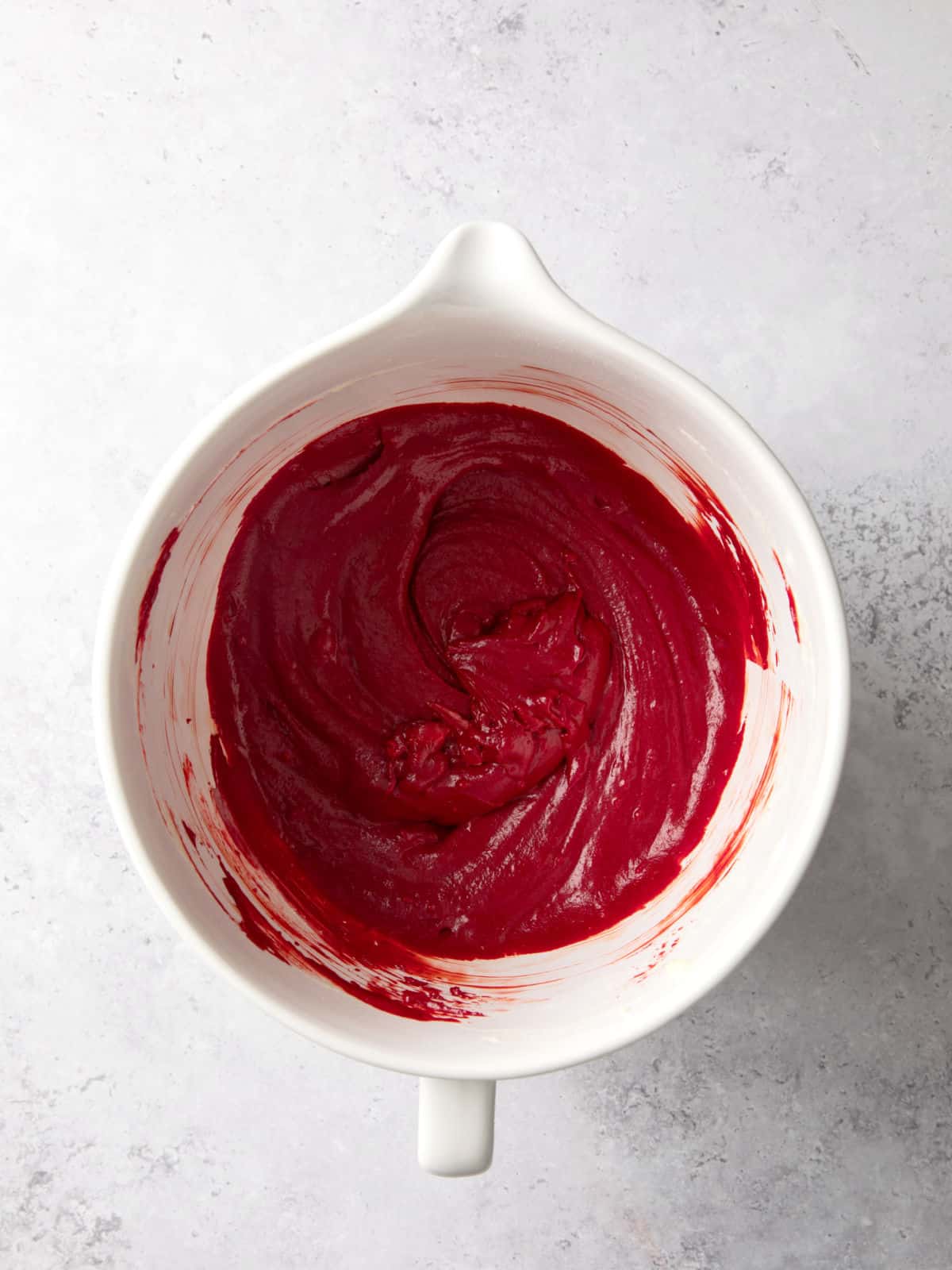 A white mixing bowl containing bright red batter on a light textured surface.