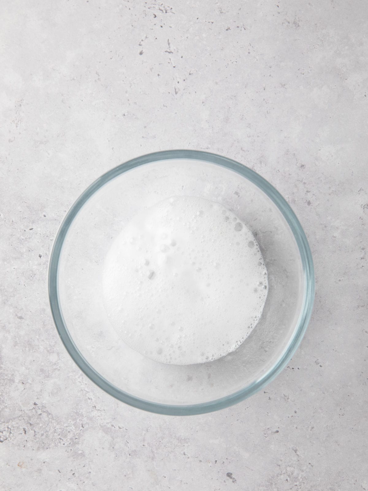 A clear glass bowl containing a foamy white liquid sits on a light gray, speckled surface.