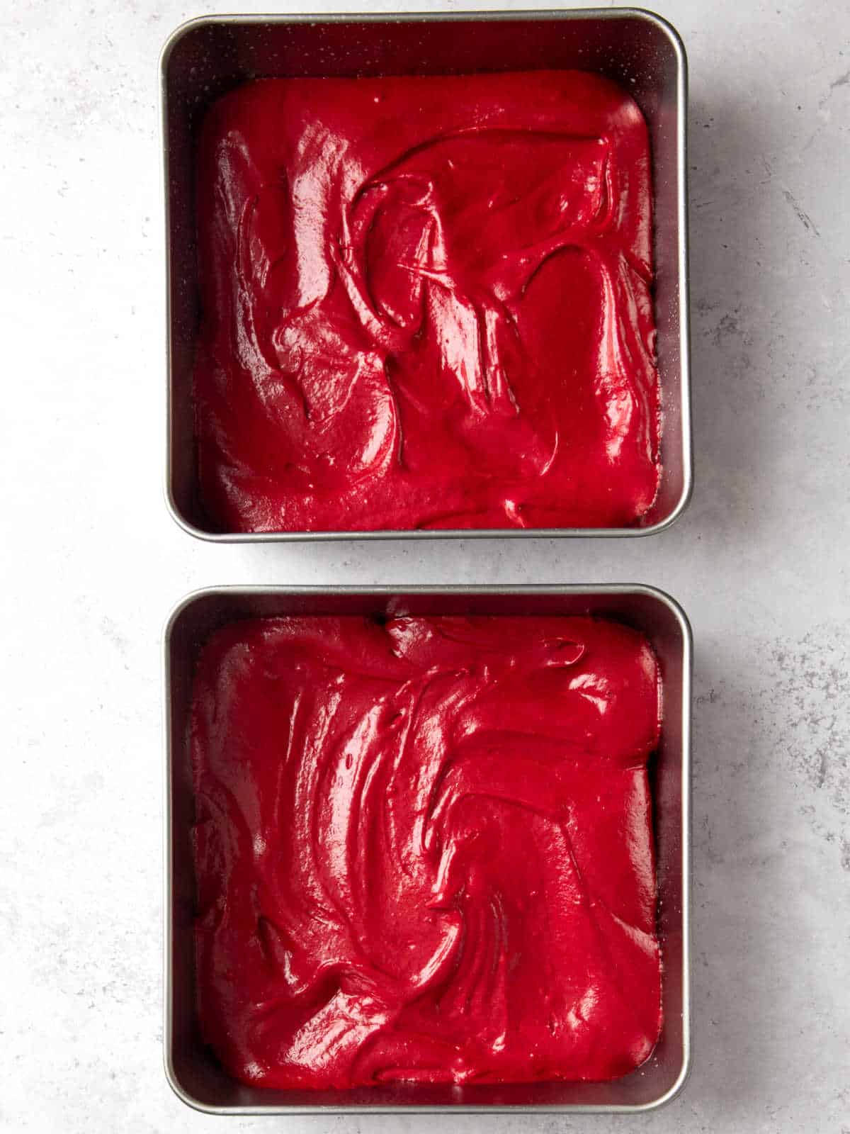 Two square baking pans filled with smooth, bright red cake batter, placed on a light marble surface.