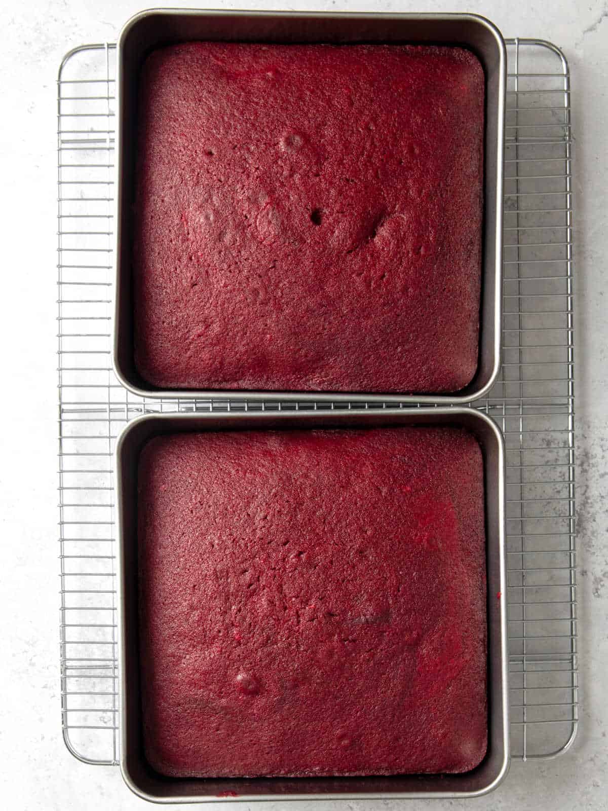 Two square red velvet cakes in metal pans cooling on a wire rack, placed on a light countertop.