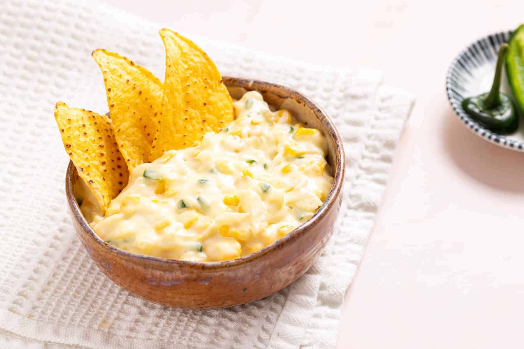 An image of jalapeno corn in a wooden bowl with tortilla chips on the side.