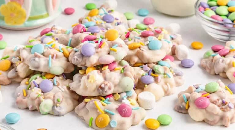 A pile of no-bake cookies with pastel candy-coated chocolates and colorful sprinkles on a white surface, with bowls of candies in the background.